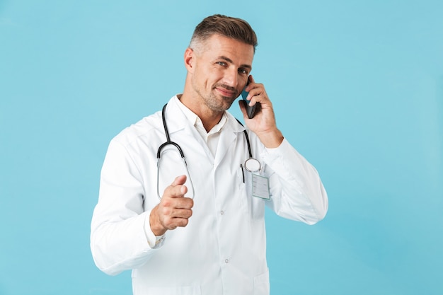 Foto de médico positivo vistiendo bata blanca y un estetoscopio hablando por teléfono celular, que se encuentran aisladas sobre la pared azul