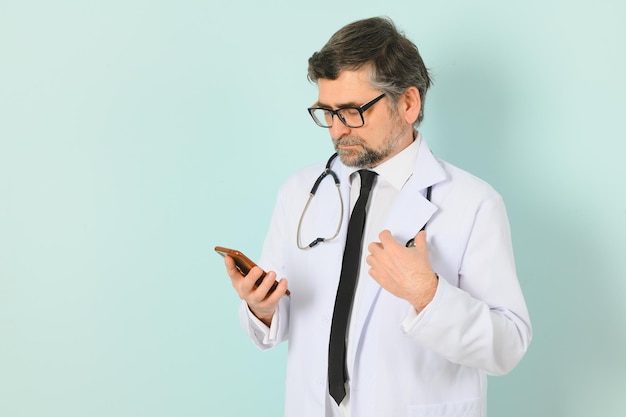 Foto de médico vistiendo bata blanca y estetoscopio sosteniendo teléfono móvil de pie aislado sobre fondo azul.