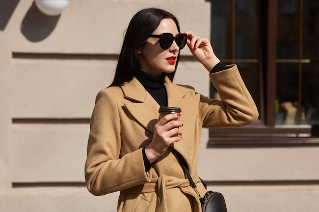 Foto de moda al aire libre de joven hermosa aislada sobre el edificio de la calle, elegante dama levantando sus gafas de sol y mirando a otro lado, disfruta bebiendo bebidas calientes para llevar mientras camina solo.