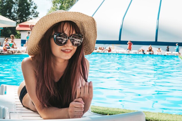 Foto de moda de chica hermosa sexy en top negro y gafas de sol relajantes acostado en un sillón blanco contra la piscina pública azul ... Retrato de estilo de vida al aire libre, Fashion Beauty Swag Girl.