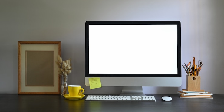 Foto foto del monitor de la computadora de la pantalla en blanco del espacio de trabajo que se coloca en el escritorio de trabajo y rodeado por un marco de imagen, un portalápices, una pila de libros, un mouse inalámbrico, un teclado, una taza de café y hierba silvestre en un jarrón.