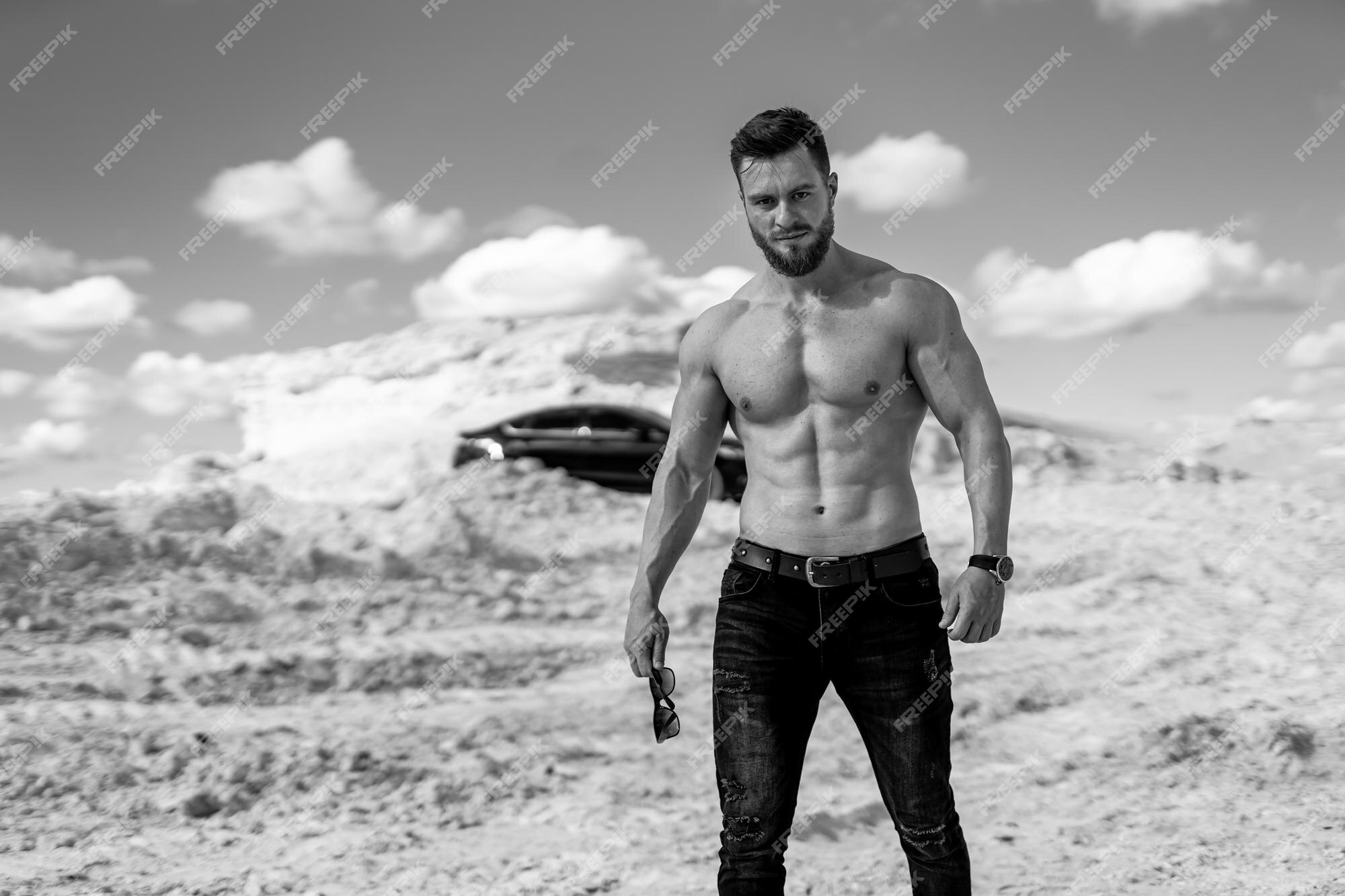 Foto monocromática de un chico guapo en montañas blancas. culturista  posando en reloj de pulsera y sosteniendo gafas de sol negras. coche  moderno en el fondo.