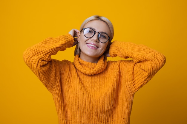 Foto monocromática de uma mulher loira com óculos tocando seu cabelo e sorrindo na parede amarela do estúdio