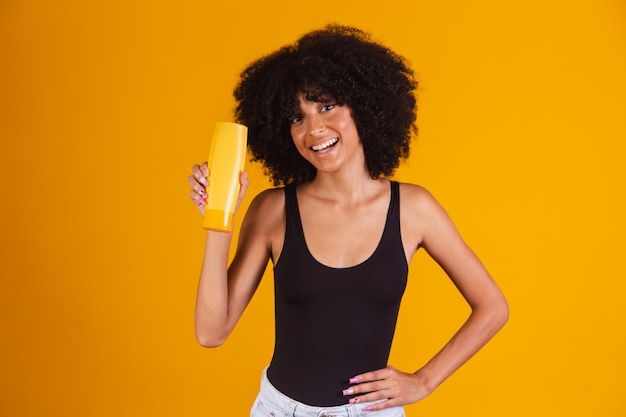 Foto de morena afro natural sostiene gel champú de botella mirando a la cámara sobre fondo amarillo