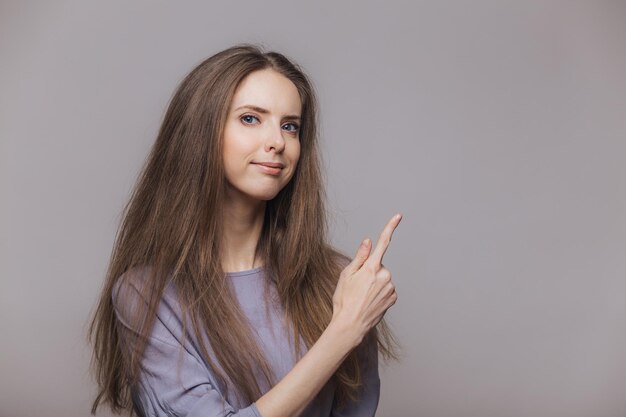 Una foto de una morena de aspecto agradable y serio con ojos azules vestida de manera informal señala con el dedo índice en un espacio de copia en blanco aislado sobre fondo gris La mujer anuncia un nuevo producto en el interior