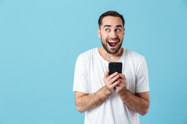 Foto de morena europea hombre vestido con camiseta básica riendo y sosteniendo smartphone aislado sobre azul