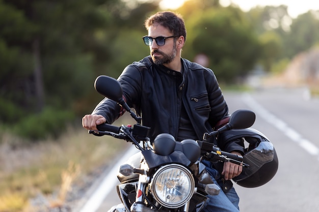 Foto de motociclista barbudo guapo mirando hacia los lados mientras está sentado en su moto y sosteniendo el casco.