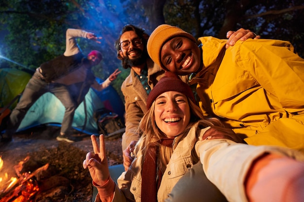 Foto foto móvil de amigos multirraciales sonriendo y tomando selfies en el bosque acampando por la noche