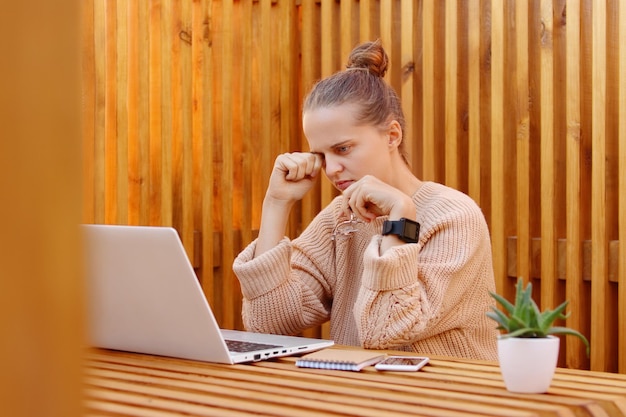 Foto de una mujer adulta joven exhausta y atractiva con un peinado de moño que usa un suéter beige que trabaja en una computadora portátil contra una pared de madera con exceso de trabajo frotándose los ojos y siente dolor en los ojos