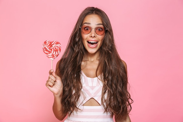 Foto de mujer alegre de 20 años con gafas de sol riendo y sosteniendo piruleta, aislado sobre pared rosa