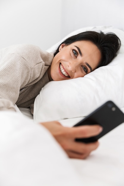 Foto de mujer alegre de 30 años con smartphone, mientras está acostado en la cama con ropa blanca en casa