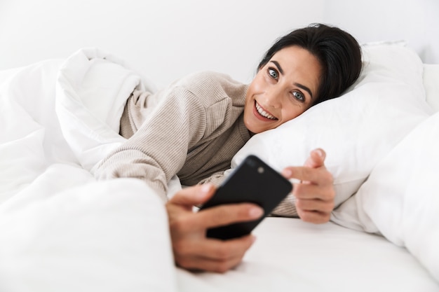 Foto de mujer alegre de 30 años con smartphone, mientras está acostado en la cama con ropa blanca en casa