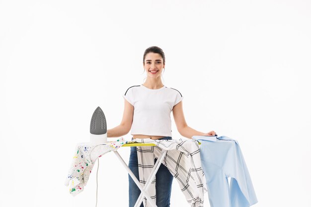 Foto de mujer alegre ama de casa de 20 años vistiendo pantalones vaqueros casuales planchando ropa limpia a bordo aislado sobre pared blanca