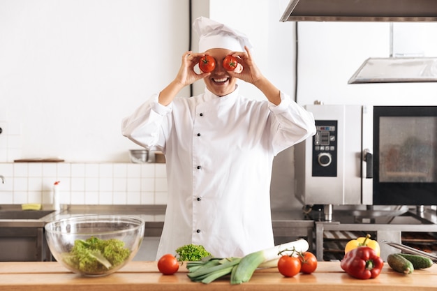 Foto de mujer alegre chef vistiendo uniforme blanco cocinar comida con verduras frescas, en la cocina del restaurante