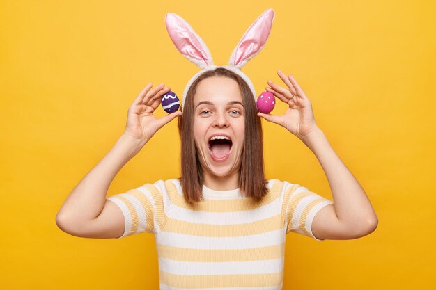 Foto de una mujer alegre y emocionada que usa orejas de conejo sosteniendo coloridos huevos de Pascua divirtiéndose mientras celebra gritando con cara feliz aislada en un fondo amarillo