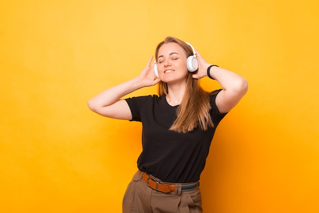 Foto de mujer alegre escuchando música en auriculares inalámbricos blancos sobre fondo amarillo