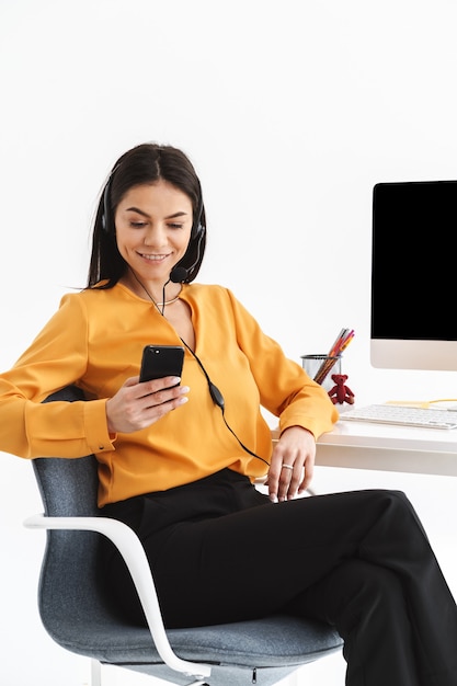 Foto de mujer alegre operador de teléfono con auriculares con micrófono sosteniendo el teléfono inteligente y hablando con el cliente mientras trabaja en la oficina