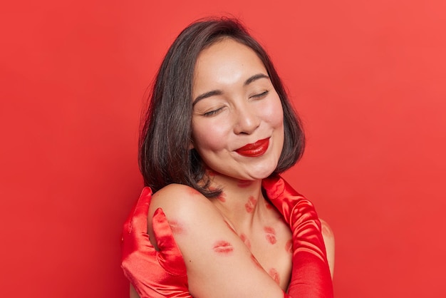 Foto de mujer asiática relajada con cabello oscuro abraza su propio cuerpo aplica lápiz labial rojo tiene cuerpo desnudo con rastros de labios aislados sobre fondo rojo sueña con algo. concepto de amor propio