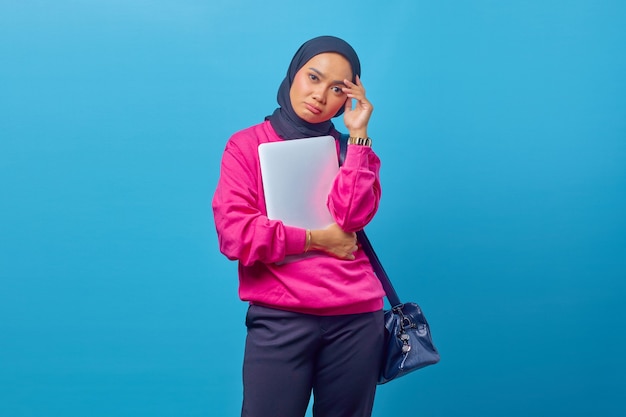 Foto de mujer asiática triste vistiendo chaqueta rosa llevando portátil mirando hacia adelante con las manos sosteniendo la cabeza aislado sobre fondo azul.