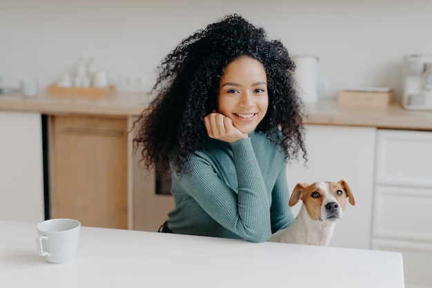 La foto de una mujer de aspecto agradable vestida con ropa informal tiene poses de cabello tupido y rizado contra el interior de la cocina con un perro pedigrí que tiene café matutino y disfruta de sonrisas agradables de fin de semana en la cámara