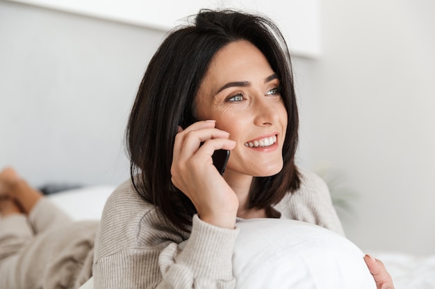 Foto de mujer atractiva de 30 años mediante teléfono móvil, mientras está acostado en la cama con ropa blanca en una habitación luminosa