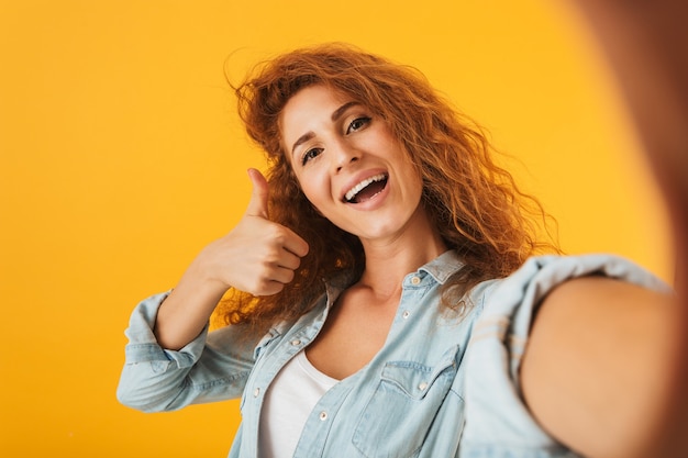 Foto de mujer atractiva rizada sonriendo y mostrando el pulgar hacia arriba mientras toma una foto selfie, aislado sobre fondo amarillo