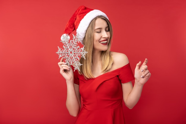 Foto de mujer atractiva con sombrero de Navidad y vestido rojo, juega con juguetes de Navidad y sonrisa. Aislado sobre fondo rojo.