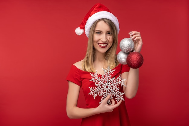 Foto foto de mujer atractiva con sombrero de navidad y vestido rojo, tiene juguetes de navidad y sonrisa feliz. aislado sobre fondo rojo.