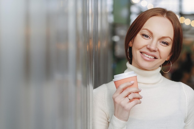 La foto de una mujer atractiva con una sonrisa con dientes tiene maquillaje vestido con un suéter de cuello alto blanco con cuello sostiene café para llevar disfruta del tiempo libre expresa buenas emociones Concepto de personas y ocio