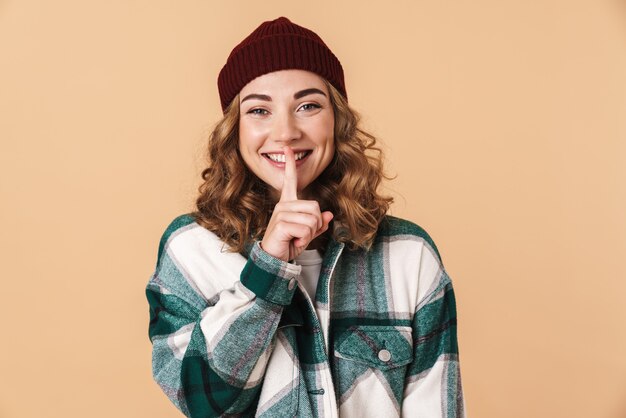 Foto de mujer bastante alegre en gorro de lana haciendo gesto de silencio y sonriendo aislado en beige