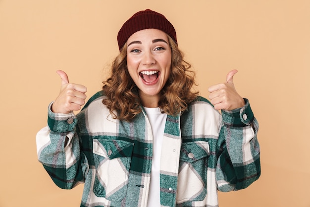 Foto de mujer bastante alegre en gorro de lana riendo y gesticulando Thumbs up aislado en beige