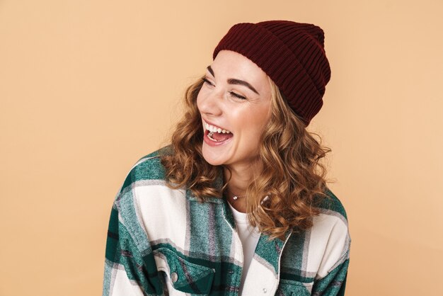 Foto de mujer bastante alegre en gorro de lana riendo y mirando a un lado aislado en beige