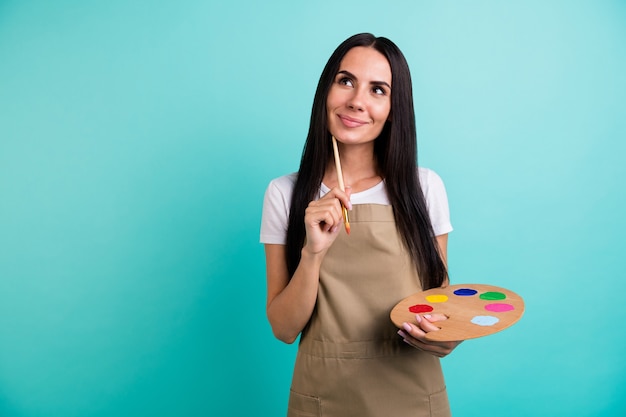 Foto de mujer beautfiul linda positiva alegre mirando hacia arriba en un ataque de pensamientos fantaseando qué pintar sosteniendo pincel paleta de colores aislado fondo vivo de color verde azulado