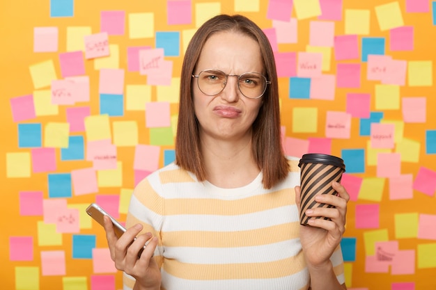 Foto de una mujer de cabello castaño triste y deprimida que usa camiseta posando contra notas adhesivas coloridas en una pared amarilla bebiendo café usando el teléfono móvil con la cara fruncida y mirando la cámara