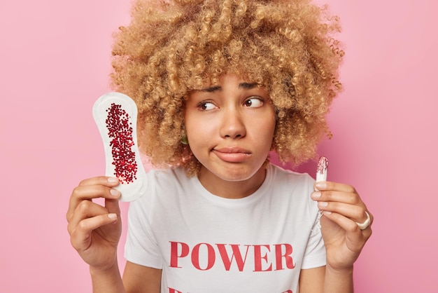 La foto de una mujer de cabello rizado disgustada habla sobre el sangrado durante la menstruación sostiene una toalla sanitaria sucia y un tampón muestra diferentes tipos de productos de higiene femenina aislados en una pared rosa