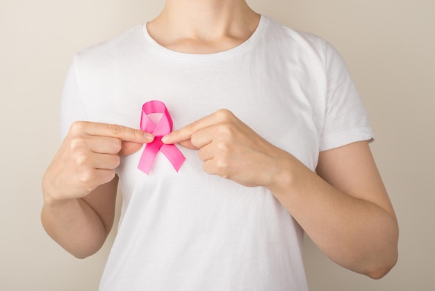 Foto de una mujer con una camiseta blanca sosteniendo un símbolo de cinta rosa de concienciación sobre el cáncer de mama en un fondo gris aislado