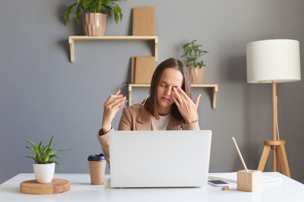 La foto de una mujer cansada y soñolienta con anteojos sentada en el lugar de trabajo con una laptop trabajando largas horas en línea siente que le duelen los ojos frotándose el ojo exhausto