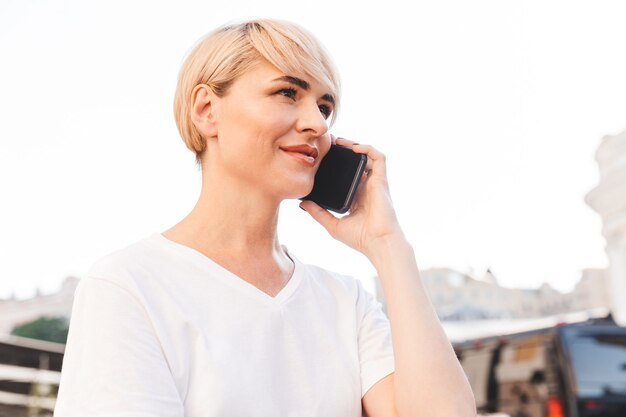 Foto de mujer caucásica alegre vistiendo camiseta blanca sentado en el café de verano al aire libre y hablando por teléfono celular