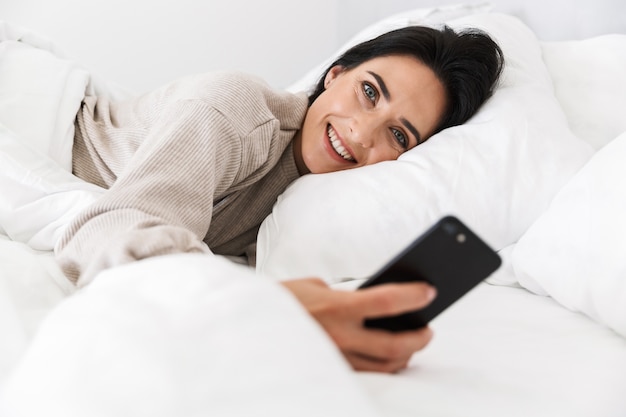 Foto de mujer complacida de 30 años con smartphone, mientras está acostada en la cama con ropa blanca en casa