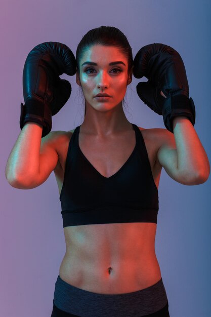 Foto de mujer delgada sexual de 20 años en ropa deportiva y guantes de boxeo negros practicando en el gimnasio, aislado sobre la pared púrpura