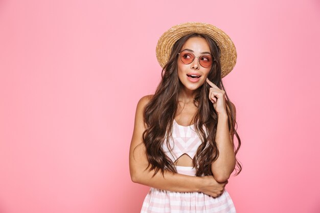 Foto de mujer elegante de 20 años con gafas de sol y sombrero de paja sonriendo, aislado sobre pared rosa