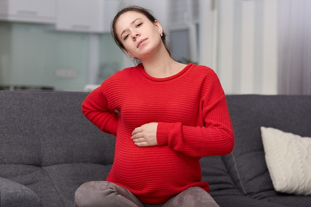 La foto de una mujer embarazada cansada disgustada siente dolor de espalda, mantiene la mano sobre el vientre hinchado, se sienta en el sofá con un cojín, usa un suéter rojo suelto