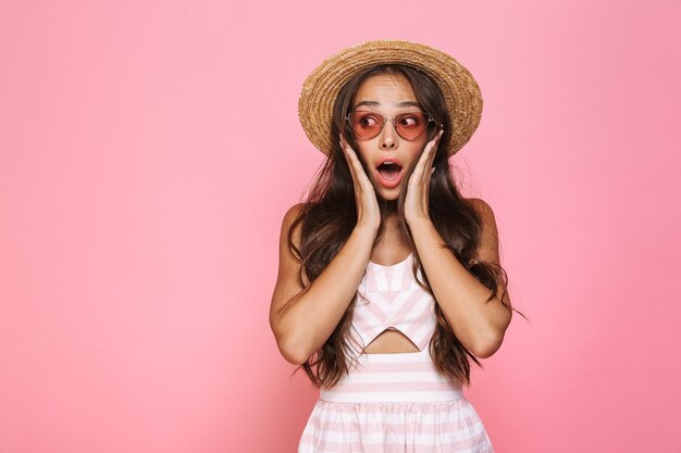 Foto de mujer emocionada de 20 años con gafas de sol y sombrero de paja gritando, aislado sobre pared rosa