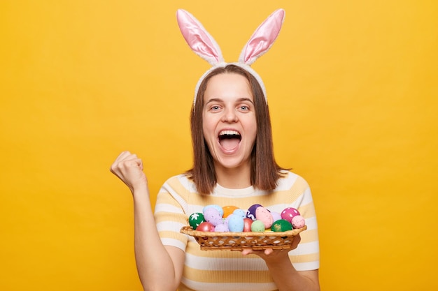 La foto de una mujer emocionada, feliz y satisfecha con orejas de conejo sosteniendo coloridos huevos de Pascua encontró el mayor número de huevos en la caza de Pascua con el puño cerrado aislado en un fondo amarillo