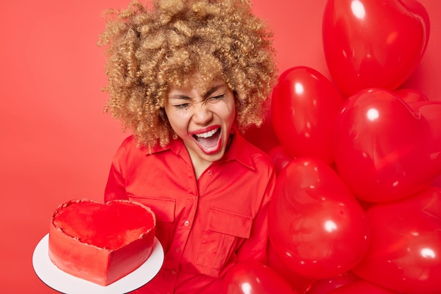 Foto de una mujer emocional disgustada con el pelo rizado sostiene un pastel dulce preparado para un montón de globos de helio celebra el cumpleaños viene en una fiesta aislada sobre fondo rojo Celebración