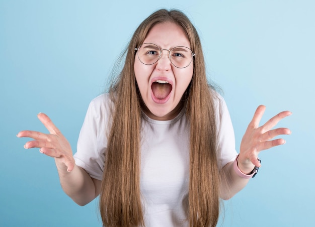 Foto de mujer enojada con gafas en ropa casual gritando de ira, cabello despeinado, aislado sobre fondo azul.