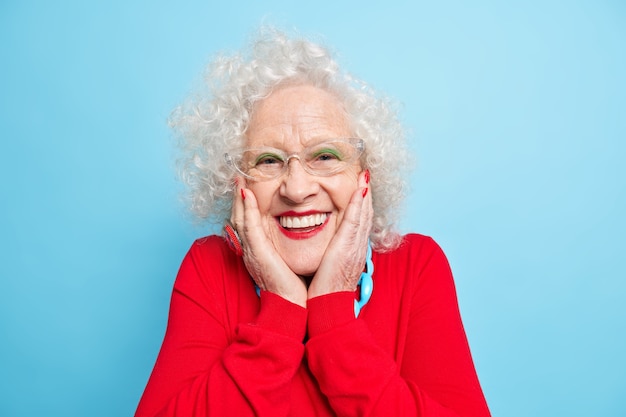 Foto foto de mujer europea de pelo gris positivo arrugado mantiene las manos en las mejillas sonríe agradablemente