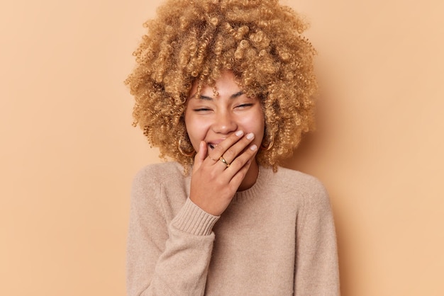 La foto de la mujer europea de pelo rizado llena de alegría disfruta viendo algo divertido cubre la boca con la mano se ríe felizmente trata de ocultar las emociones viste un jersey casual aislado sobre fondo beige