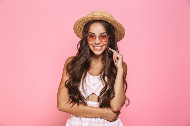 Foto de mujer feliz 20s con gafas de sol y sombrero de paja sonriendo, aislado sobre pared rosa