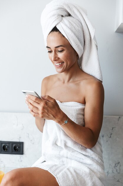 Foto de una mujer feliz sonriente alegre joven en toalla después de la ducha del procedimiento del balneario adentro en casa en la cocina usando el teléfono móvil.